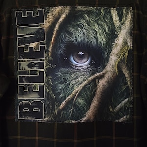 May include: Dark green and brown plaid flannel shirt with a large patch on the back. The patch features a detailed eye peering through a forest, with the word "BELIEVE" printed vertically in white.