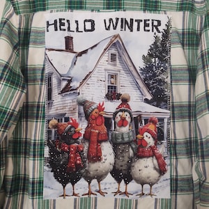 May include: A green and white plaid flannel shirt with a patch featuring four chickens wearing winter hats and scarves in front of a snow-covered house. The text "HELLO WINTER" is above the image.