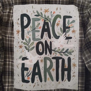 May include: A green and brown plaid flannel shirt with a hand-painted design on the back. The design features the words "PEACE ON EARTH" in a mix of green and black lettering, with illustrations of doves and foliage.