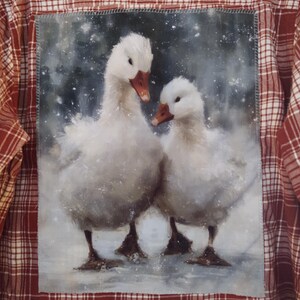 May include: Red and white plaid flannel shirt with a rectangular patch featuring a painting of two white geese in a snowy scene. The geese have orange beaks and stand close together.