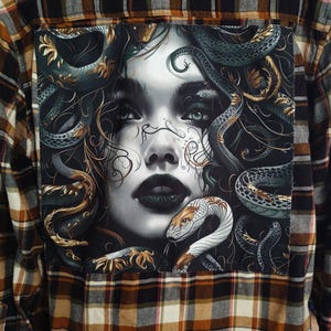 May include: A brown, black, and white plaid flannel shirt with a Medusa graphic on the back. The graphic features a grayscale portrait of a woman with snakes for hair, with green and gold accents.