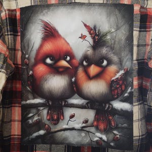 May include: Plaid flannel shirt with a large graphic of two cartoon birds perched on a snowy branch. The birds are red and gray with detailed feathers and are set against a wintery background. The shirt has a classic red, black, and white plaid pattern.