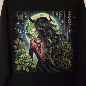 May include: Black long-sleeve shirt with a green, blue, and white graphic print of a woman with horns and a tattoo of the Taurus zodiac symbol. The graphic also includes green roses and a crescent moon.