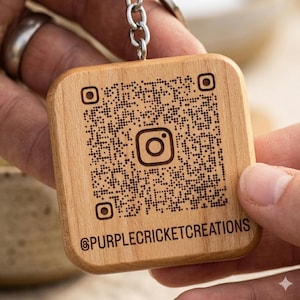 May include: Wooden square keychain featuring a QR code and the Instagram logo. The keychain has rounded corners and a silver chain. The text "@PURPLECRICKETCREATIONS" is engraved at the bottom. The wood has a natural grain.