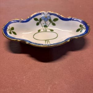 May include: An oval porcelain dish with a scalloped edge, featuring a white interior painted with a single blue flower, green leaves, and a green oval outline. The rim is decorated with a bold blue border and a thin, worn gold-colored trim.
