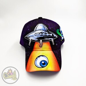 Puede incluir: Una gorra de béisbol morada con un gráfico de OVNI en blanco y negro y un gráfico de ojo azul en la parte delantera. El OVNI emite un rayo de luz amarillo.