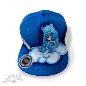 May include: A blue baseball cap featuring a white cloud with a blue Care Bear sitting on it. The Care Bear is frowning and holding a heart. The cap has a white star pattern.