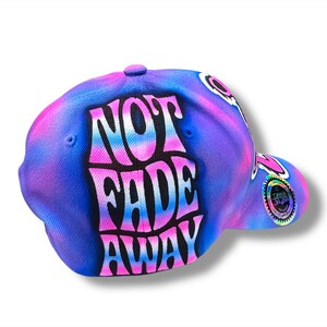 Puede incluir: Gorra de b&eacute;isbol con un dise&ntilde;o tie-dye en tonos azules, morados y rosas. La gorra presenta las palabras "NOT FADE AWAY" en una fuente audaz y estilizada. Un emblema circular es visible en el lateral.
