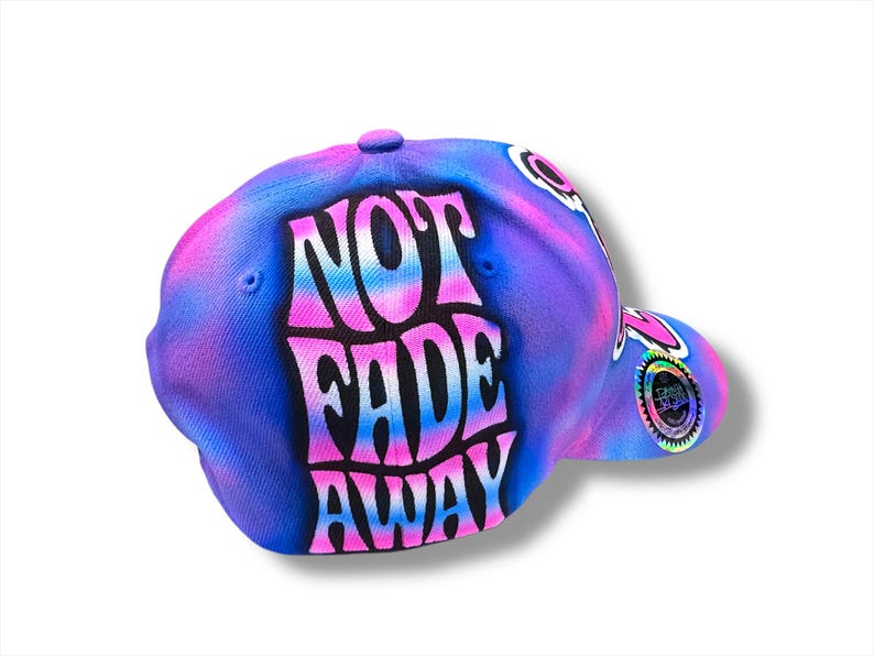 Puede incluir: Gorra de b&eacute;isbol con un dise&ntilde;o tie-dye en tonos azules, morados y rosas. La gorra presenta las palabras "NOT FADE AWAY" en una fuente audaz y estilizada. Un emblema circular es visible en el lateral.