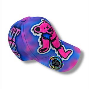 Puede incluir: Gorra de b&eacute;isbol con un dise&ntilde;o vibrante. La gorra presenta un gr&aacute;fico de oso rosa y negro sobre un fondo te&ntilde;ido anudado morado y azul. El texto "LOVE OF LIFE" es visible en el lateral. Una pegatina hologr&aacute;fica est&aacute; en la visera.