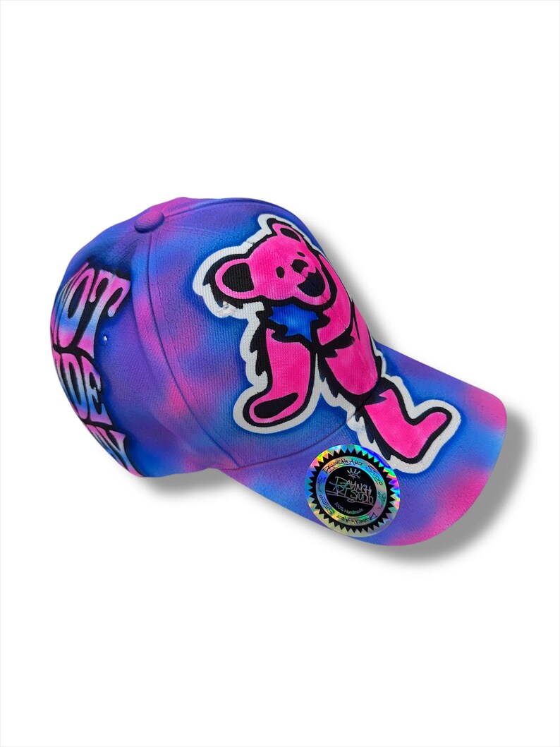 Puede incluir: Gorra de b&eacute;isbol con un dise&ntilde;o vibrante. La gorra presenta un gr&aacute;fico de oso rosa y negro sobre un fondo te&ntilde;ido anudado morado y azul. El texto "LOVE OF LIFE" es visible en el lateral. Una pegatina hologr&aacute;fica est&aacute; en la visera.