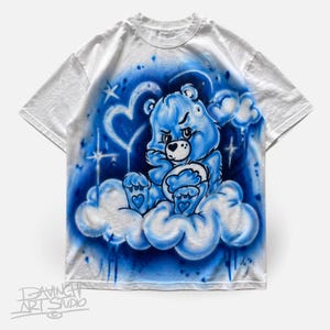 May include: White t-shirt featuring a blue Care Bear character on a cloud, with a blue and white airbrushed background. The design includes hearts, stars, and the text "DAVINGH ART STUDIO" at the bottom. The bear has an angry expression.