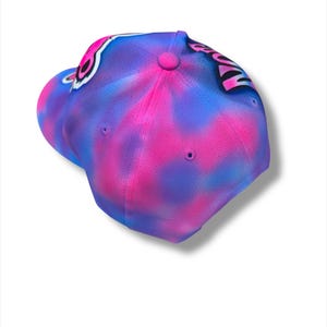 Puede incluir: Gorra de b&eacute;isbol con un vibrante dise&ntilde;o tie-dye en tonos rosa y azul. La gorra presenta una visera curva y un bot&oacute;n en la parte superior. El dise&ntilde;o incluye un elemento gr&aacute;fico en blanco y negro.