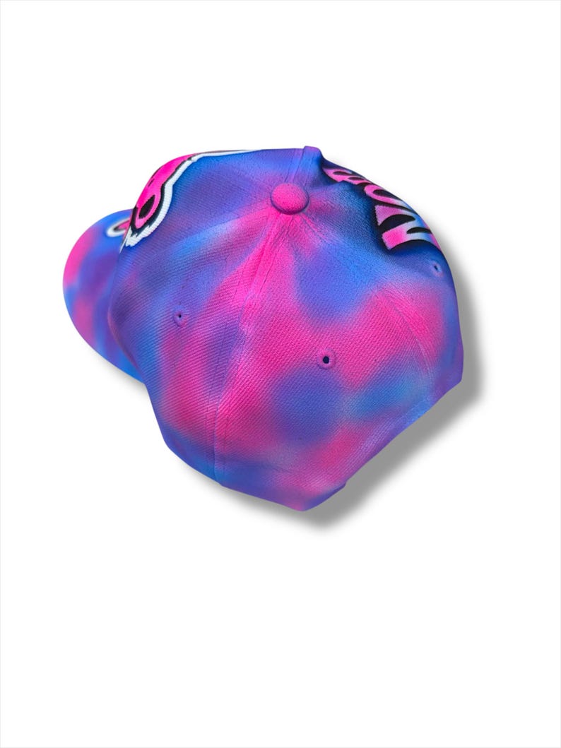 Puede incluir: Gorra de b&eacute;isbol con un vibrante dise&ntilde;o tie-dye en tonos rosa y azul. La gorra presenta una visera curva y un bot&oacute;n en la parte superior. El dise&ntilde;o incluye un elemento gr&aacute;fico en blanco y negro.