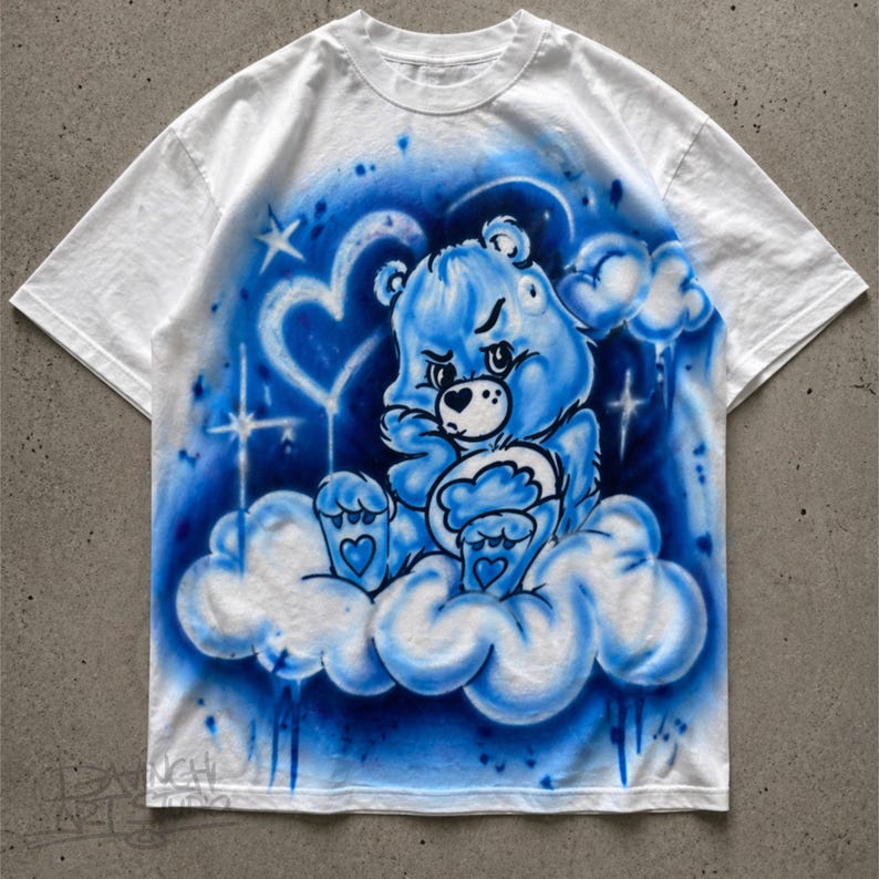 May include: White t-shirt featuring a blue cartoon bear design. The bear is sitting on a cloud, surrounded by blue accents, hearts, and stars. The design has an airbrushed effect, giving it a unique look.