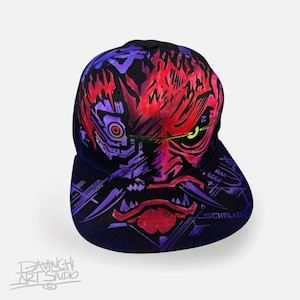 Puede incluir: Gorra de béisbol negra con un diseño gráfico estilizado y vibrante en tonos rojos, morados y rosas. El diseño incluye una ilustración con forma de rostro, la palabra "SAMURAI" y el texto "DAVINGHI ART STUDIO".