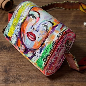 Puede incluir: Un bolso bandolera pintado a mano, colorido, con un retrato de una mujer con labios rojos y cabello rubio. El bolso tiene una correa de cuero marrón y las palabras "DAVINCI ART" son visibles. También hay un pequeño botón con texto.