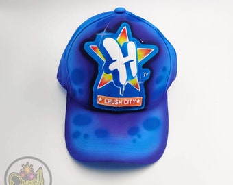 Op de afbeelding: Blauwe baseballpet met een witte ster en de tekst "Crush City" in oranje. De ster heeft een witte letter "H" erin.