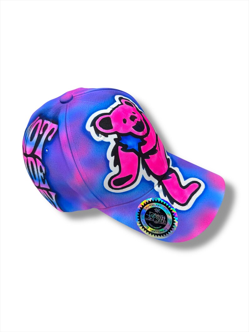 Puede incluir: Gorra de b&eacute;isbol con un vibrante dise&ntilde;o tie-dye en tonos de azul, morado y rosa. La gorra presenta un gr&aacute;fico de oso rosa y el texto "NOT DEAD" en el lateral. Una pegatina hologr&aacute;fica est&aacute; en la visera.