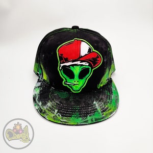 Puede incluir: Gorra de béisbol negra de ala plana con un gráfico de alienígena verde que lleva un sombrero rojo y blanco y fuma un cigarrillo. La gorra tiene un diseño de salpicaduras de pintura verde y blanca en la visera y los lados.