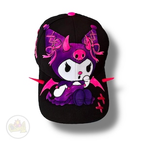 Puede incluir: Gorra de béisbol negra con un diseño de personaje de dibujos animados morado y rosa. El personaje tiene un cráneo en la cabeza, cuernos rosas y alas rosas. La gorra tiene un botón rosa en la parte superior y detalles rosas.