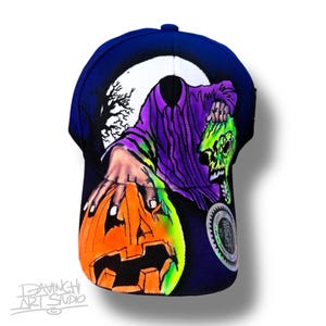 Puede incluir: Gorra de béisbol azul marino con un diseño de Halloween. El diseño incluye una figura con capucha morada sosteniendo una calavera verde y una calabaza naranja. Una luna llena y una silueta de árbol están en el fondo. El texto "DAVINGHI ART STUDIO" está en la parte inferior.