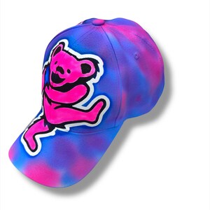Puede incluir: Gorra de b&eacute;isbol con un dise&ntilde;o tie-dye en tonos azules, morados y rosas. La gorra presenta un gr&aacute;fico de oso rosa con un contorno blanco. La gorra tiene una visera curva y un bot&oacute;n en la parte superior.