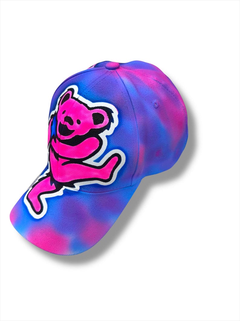 Puede incluir: Gorra de b&eacute;isbol con un dise&ntilde;o tie-dye en tonos azules, morados y rosas. La gorra presenta un gr&aacute;fico de oso rosa con un contorno blanco. La gorra tiene una visera curva y un bot&oacute;n en la parte superior.