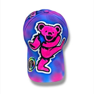 Puede incluir: Gorra de b&eacute;isbol con un dise&ntilde;o tie-dye en tonos azules y morados. Un gr&aacute;fico de un oso bailar&iacute;n rosa est&aacute; centrado en la parte delantera. La gorra tiene una visera curva y una pegatina en el lateral.