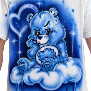 May include: White t-shirt featuring a blue cartoon bear sitting on a cloud. The bear has a grumpy expression and is surrounded by a blue and white background with stars and a heart shape. The design is airbrushed.