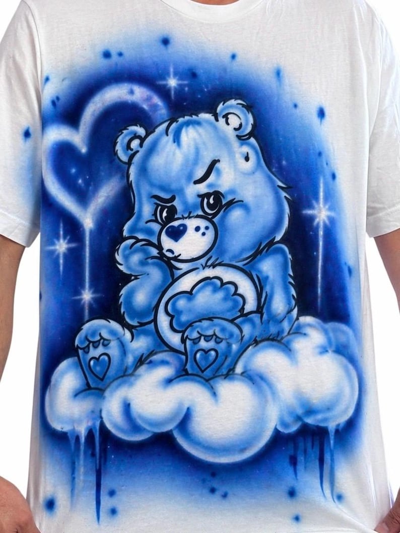 May include: White t-shirt featuring a blue cartoon bear sitting on a cloud. The bear has a grumpy expression and is surrounded by a blue and white background with stars and a heart shape. The design is airbrushed.