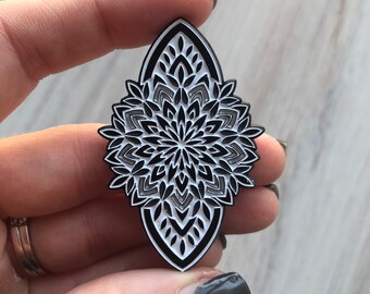 May include: Black and white enamel pin with a detailed mandala design. The pin is shaped like a teardrop and has a shiny finish.