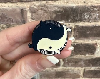 May include: A black and white enamel pin featuring two whales in a yin yang design. The whales are facing each other with their tails intertwined.