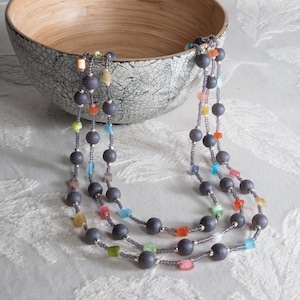 May include: Three-strand necklace featuring dark gray beads interspersed with colorful square and round beads. The necklace is displayed near a decorative wooden bowl with a cracked, gray exterior.