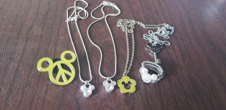 May include: Collection of silver necklaces and a ring with Mickey Mouse-themed pendants and charms. Includes a peace sign pendant, a key pendant with "BFF" text, and a ring with a Mickey Mouse design.