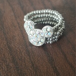 May include: Silver-toned ring featuring a Mickey Mouse head design. The Mickey Mouse head is adorned with iridescent rhinestones. The ring is made of multiple strands of small, round beads.