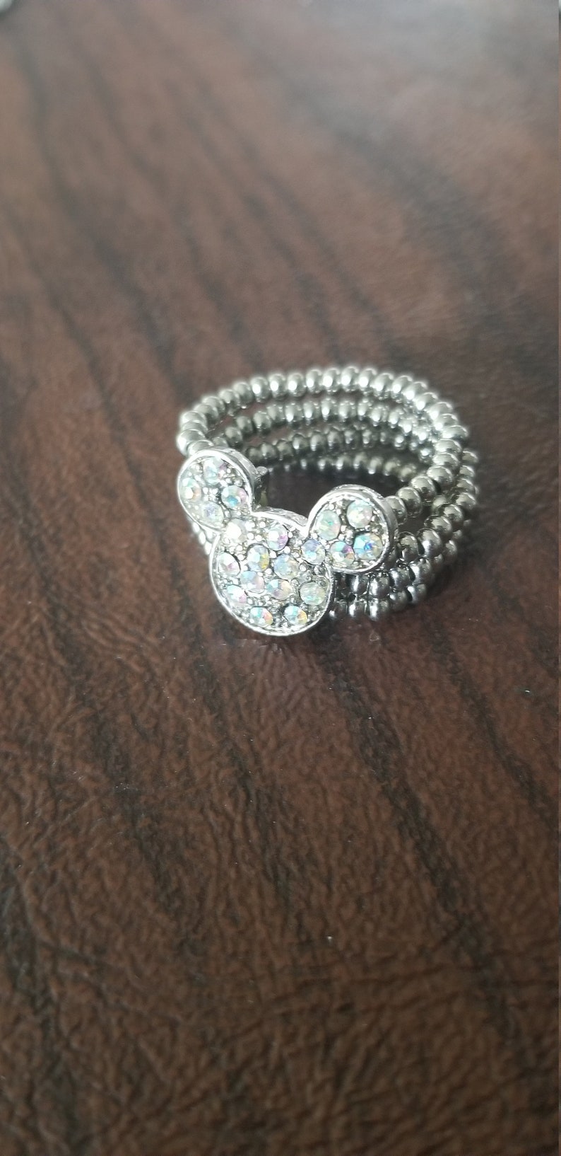 May include: Silver-toned ring featuring a Mickey Mouse head design. The Mickey Mouse head is adorned with iridescent rhinestones. The ring is made of multiple strands of small, round beads.