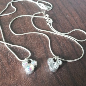 May include: Two silver necklaces with Mickey Mouse pendant charms. One charm has iridescent stones, the other has clear stones. The necklaces are displayed on a brown surface.