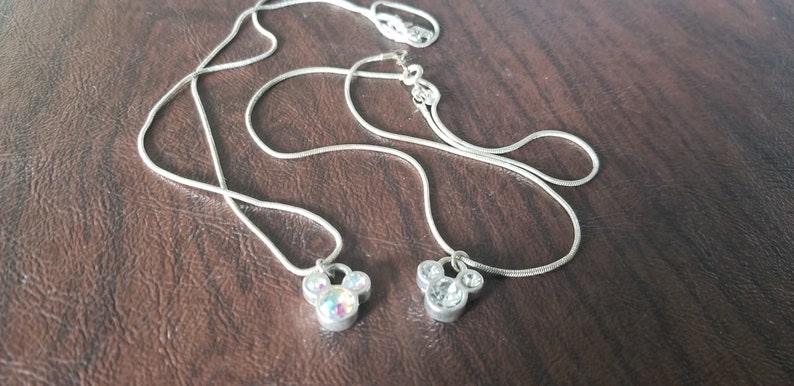May include: Two silver necklaces with Mickey Mouse pendant charms. One charm has iridescent stones, the other has clear stones. The necklaces are displayed on a brown surface.
