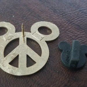 May include: A gold-colored pin shaped like Mickey Mouse ears with a peace sign in the center. The pin has a long, sharp needle for attachment and a black rubber backing piece. The pin is a Disney product.
