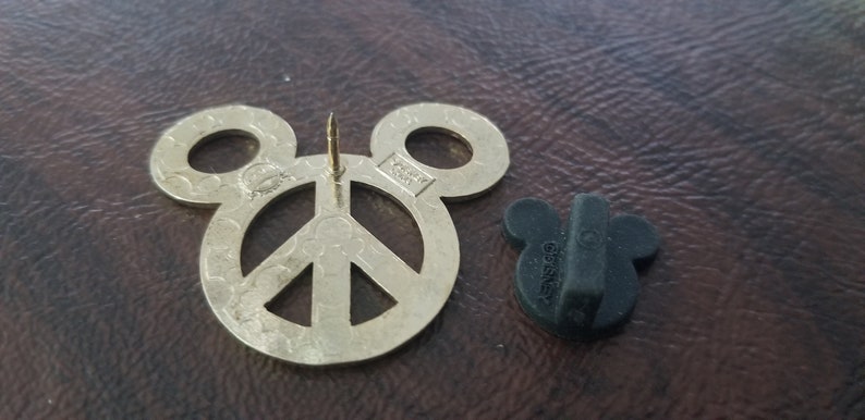 May include: A gold-colored pin shaped like Mickey Mouse ears with a peace sign in the center. The pin has a long, sharp needle for attachment and a black rubber backing piece. The pin is a Disney product.