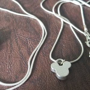 May include: Silver-toned necklaces featuring Mickey Mouse-shaped pendants. The necklaces have a delicate chain design and a lobster claw clasp. The pendants are a solid, three-dimensional design. The necklaces are displayed on a brown surface.