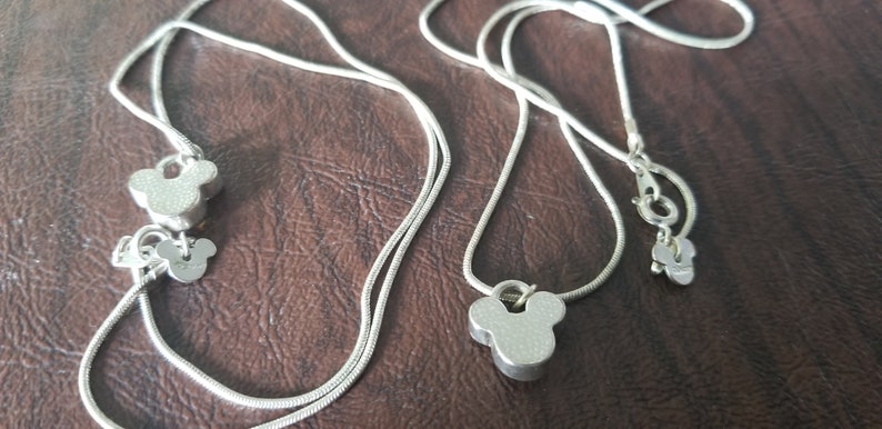 May include: Silver-toned necklaces featuring Mickey Mouse-shaped pendants. The necklaces have a delicate chain design and a lobster claw clasp. The pendants are a solid, three-dimensional design. The necklaces are displayed on a brown surface.