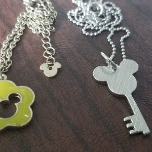 May include: Two silver necklaces with pendants. One pendant is a lime green flower shape with a Mickey Mouse silhouette. The other is a key shape with a Mickey Mouse head. Both necklaces have silver chains.