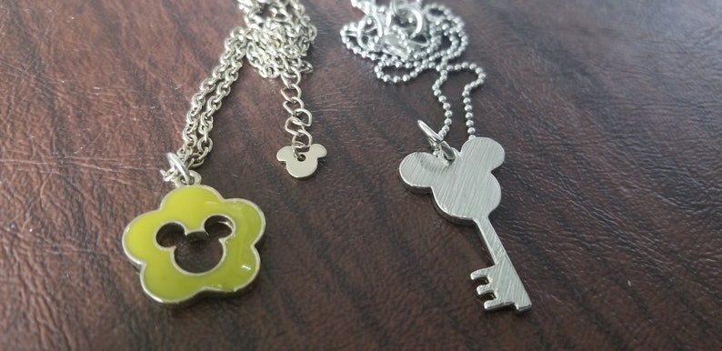 May include: Two silver necklaces with pendants. One pendant is a lime green flower shape with a Mickey Mouse silhouette. The other is a key shape with a Mickey Mouse head. Both necklaces have silver chains.