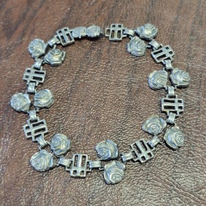 May include: A silver necklace with a repeating pattern of stylized roses and geometric shapes. The necklace is made of metal and has a delicate, feminine design.