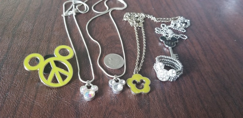 May include: A collection of silver-toned necklaces and a ring with Mickey Mouse-themed pendants and charms. One pendant is a yellow Mickey Mouse head with a peace sign. The ring and some pendants are embellished with rhinestones.