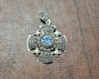 May include: Silver-toned cross pendant with an ornate design. The pendant features a central, light blue gemstone and detailed filigree work. The cross has a loop at the top for a chain. The pendant is likely made of metal.