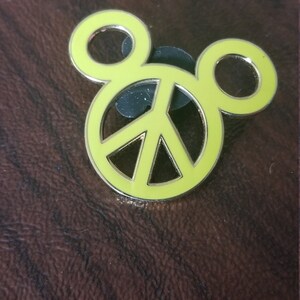 May include: A yellow enamel pin in the shape of Mickey Mouse's head, with a peace sign in the center. The pin has a silver border and a black backing. The pin is on a brown, textured surface.