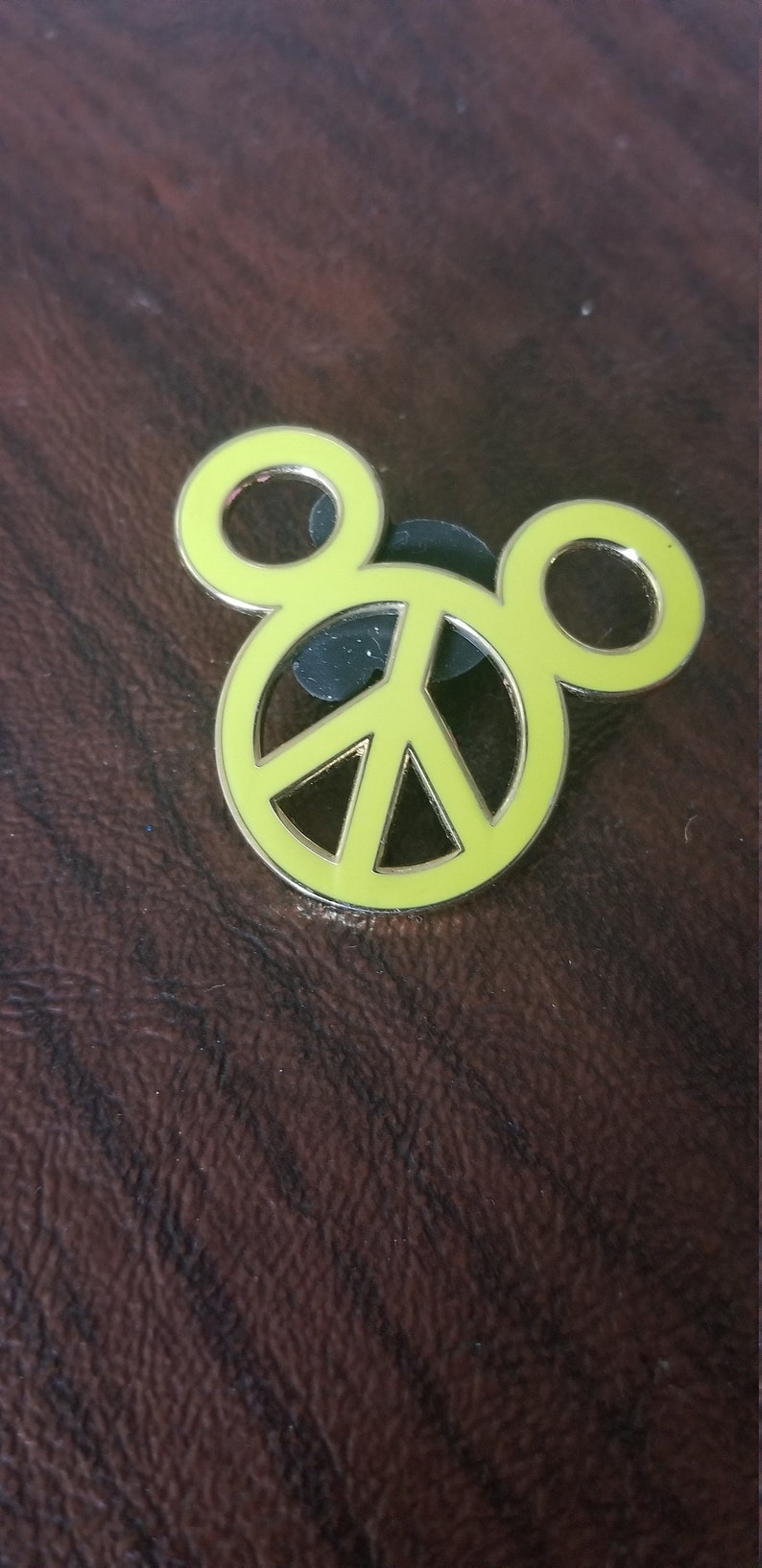 May include: A yellow enamel pin in the shape of Mickey Mouse's head, with a peace sign in the center. The pin has a silver border and a black backing. The pin is on a brown, textured surface.